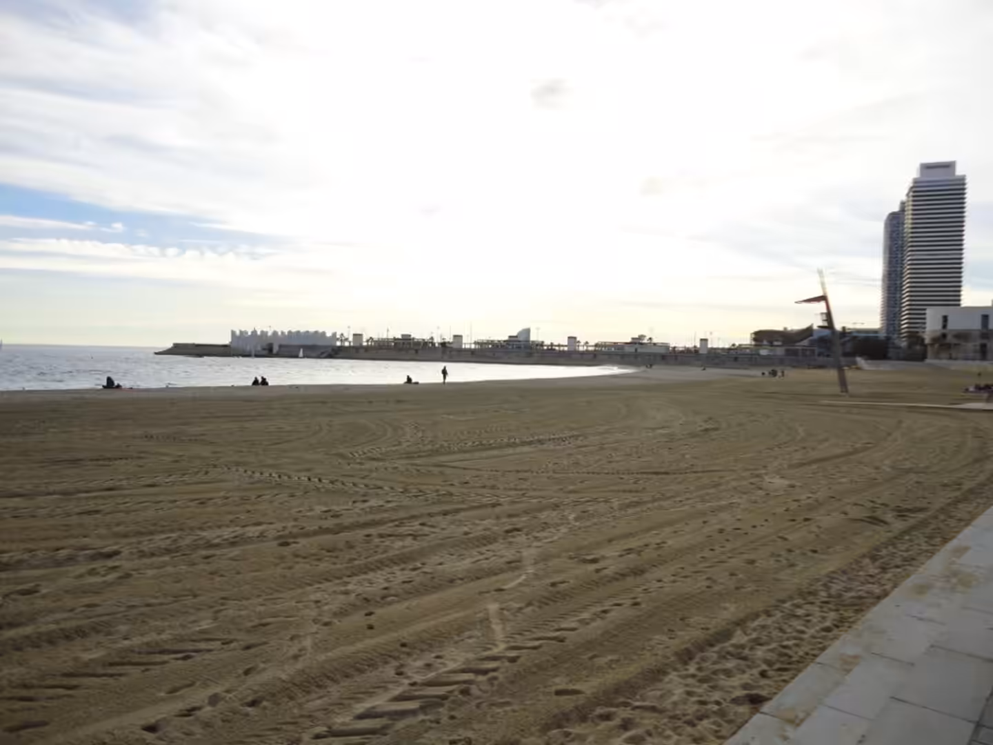 Broad sandy beach with tire marks, a calm sea, a few people walking near the water, and tall modern buildings in the distance in Barcelona.