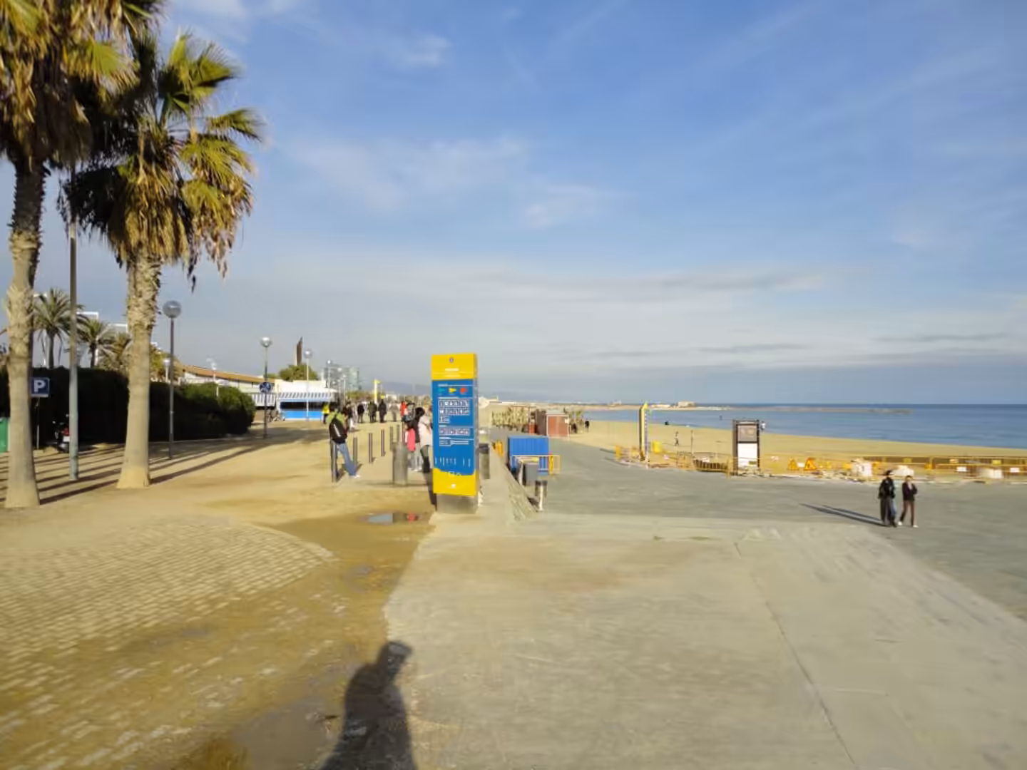 Palm-lined seaside promenade with pedestrians, beach access signage, and the Mediterranean Sea along Barceloneta Beach in Barcelona.