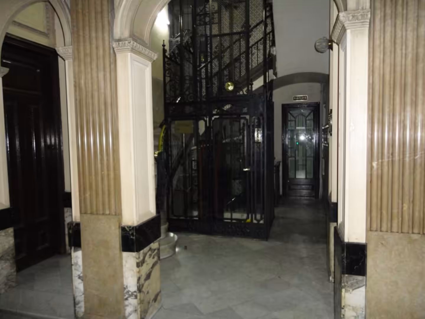 Interior of a historic apartment building with marble columns, arched doorways, and an old wrought-iron elevator cage.