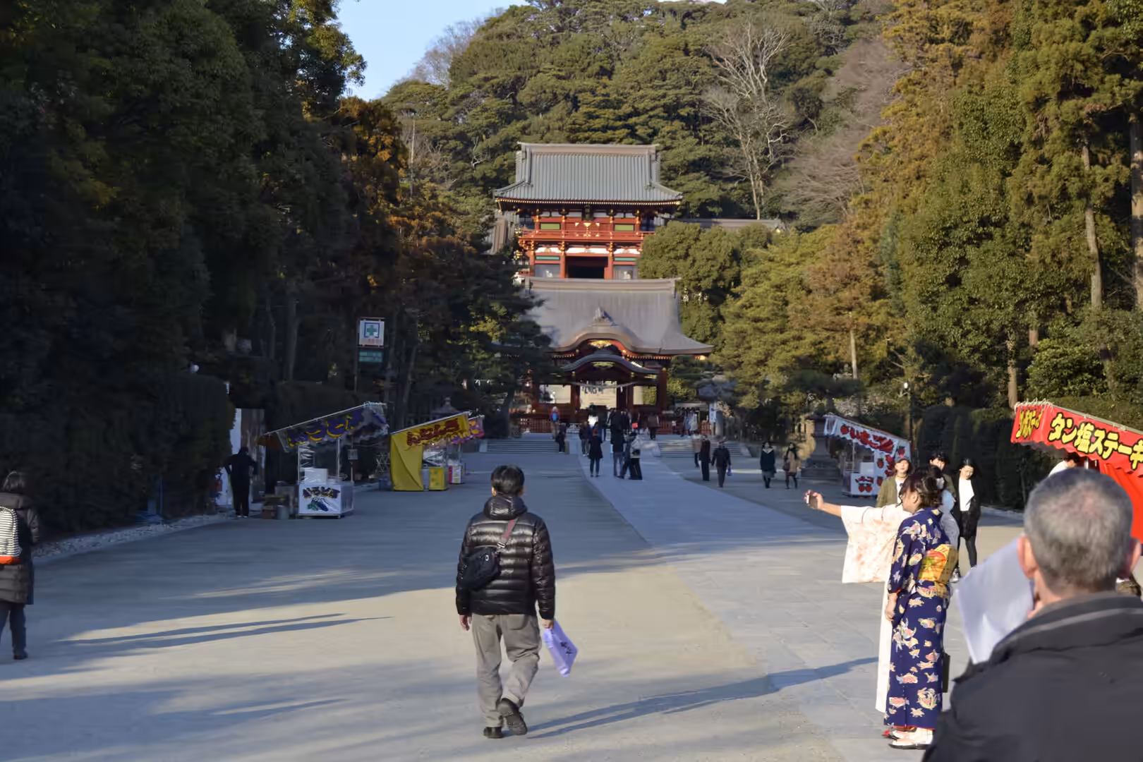 A broad gravel path runs through trees toward a large red temple gate, with visitors strolling toward the entrance.