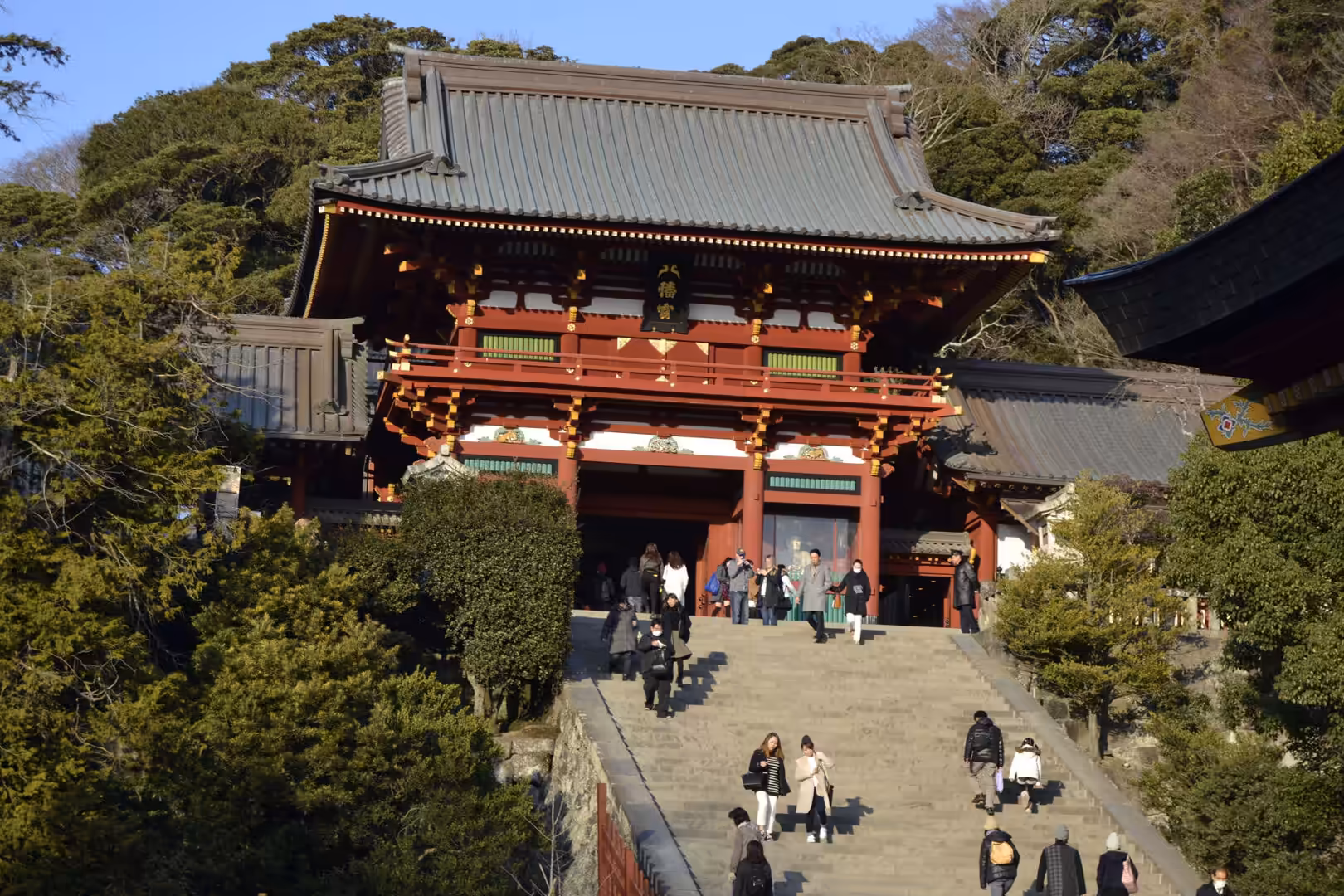 A large vermilion gate stands at the top of a wide stone staircase surrounded by greenery and visitors.
