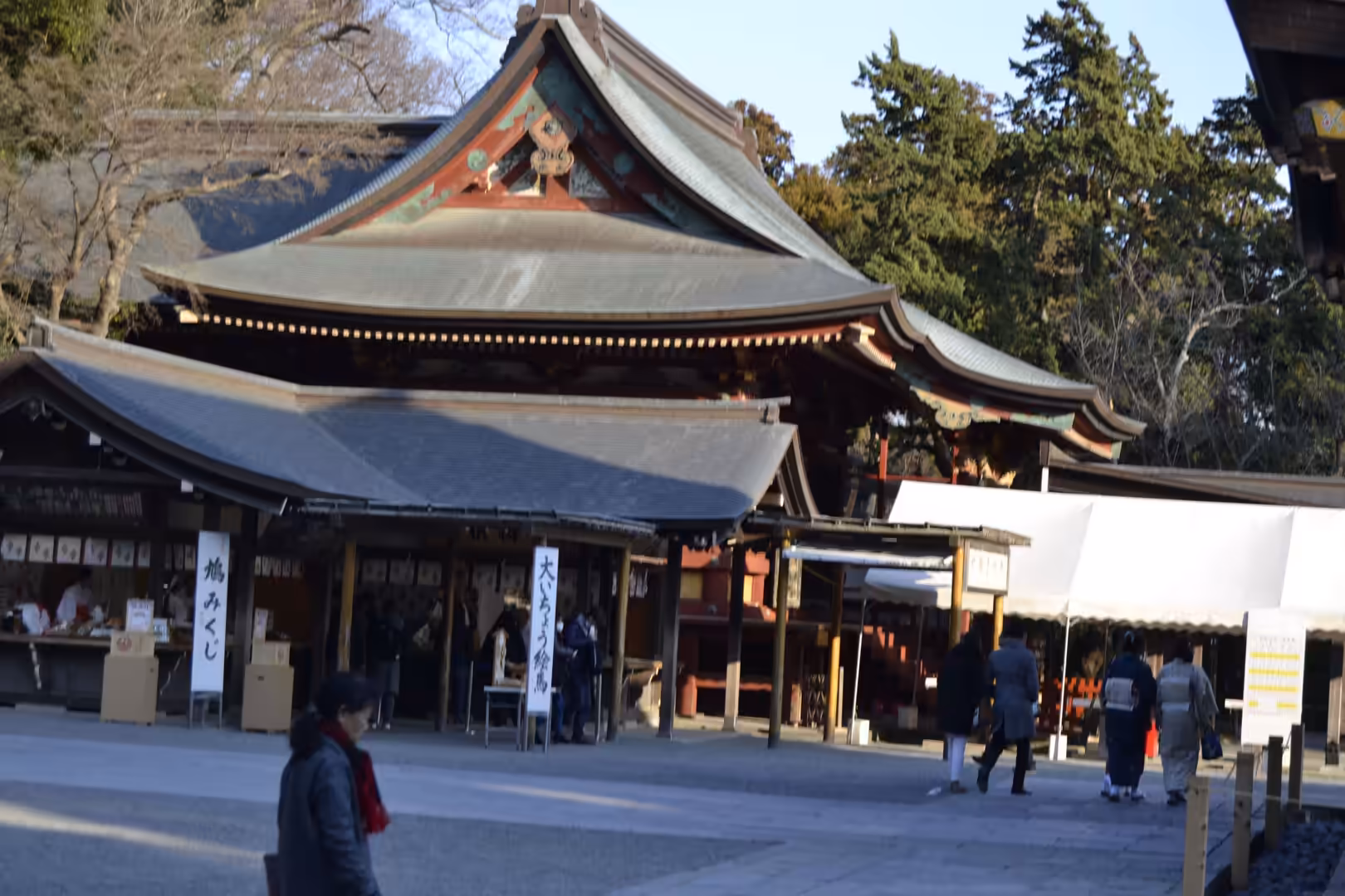 A grand wooden hall with an ornate dark tiled roof faces a courtyard where visitors browse stalls and signboards.