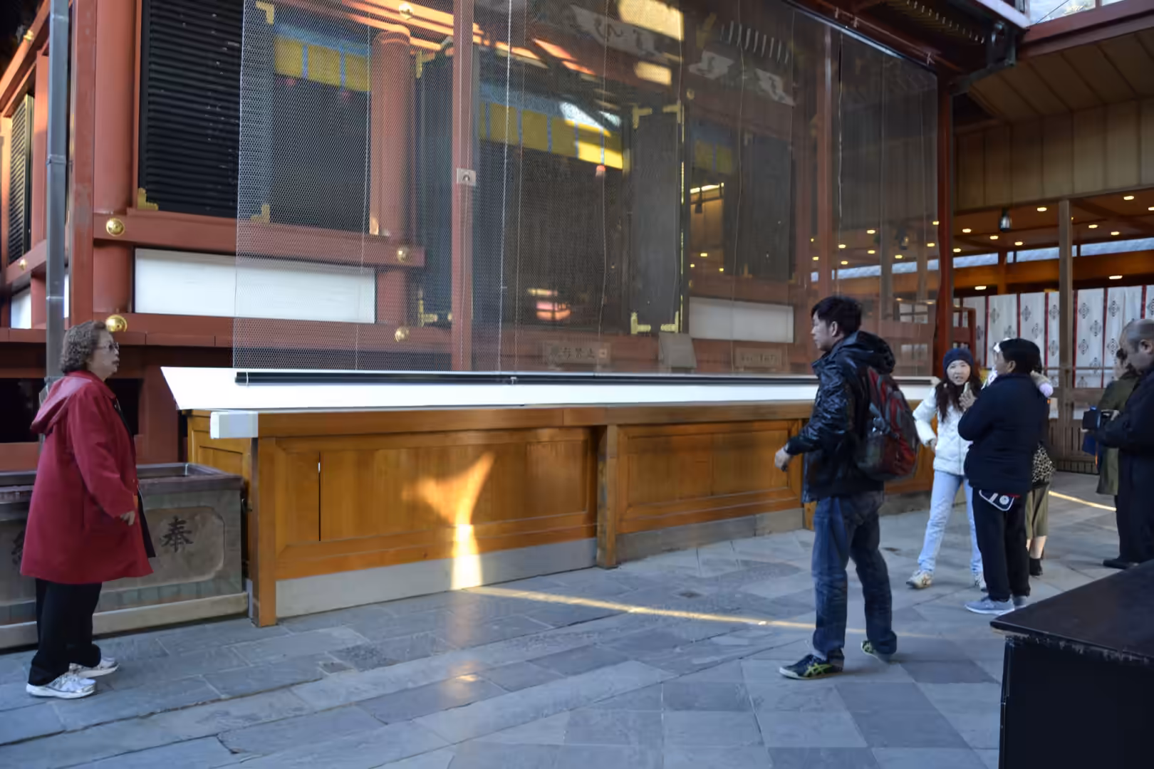 Visitors stand before a red-painted temple hall and a wooden offering box.