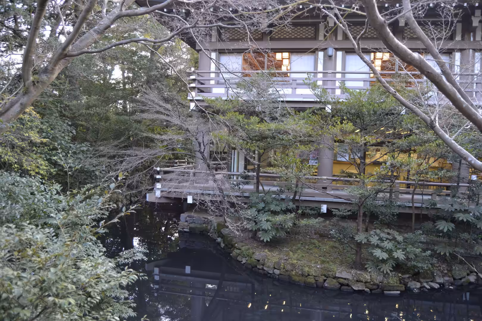 A Kamakura garden with a reflective pond, wooden bridge and a railed building behind trees.