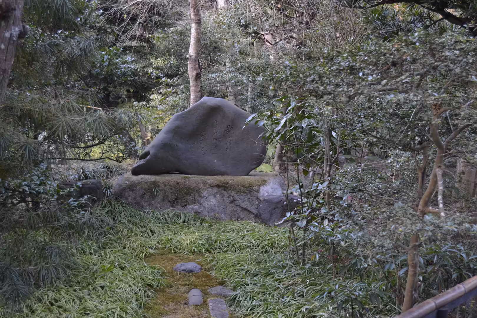 A dark moss-covered boulder rests on a stone base amid ferns in a shaded park garden.