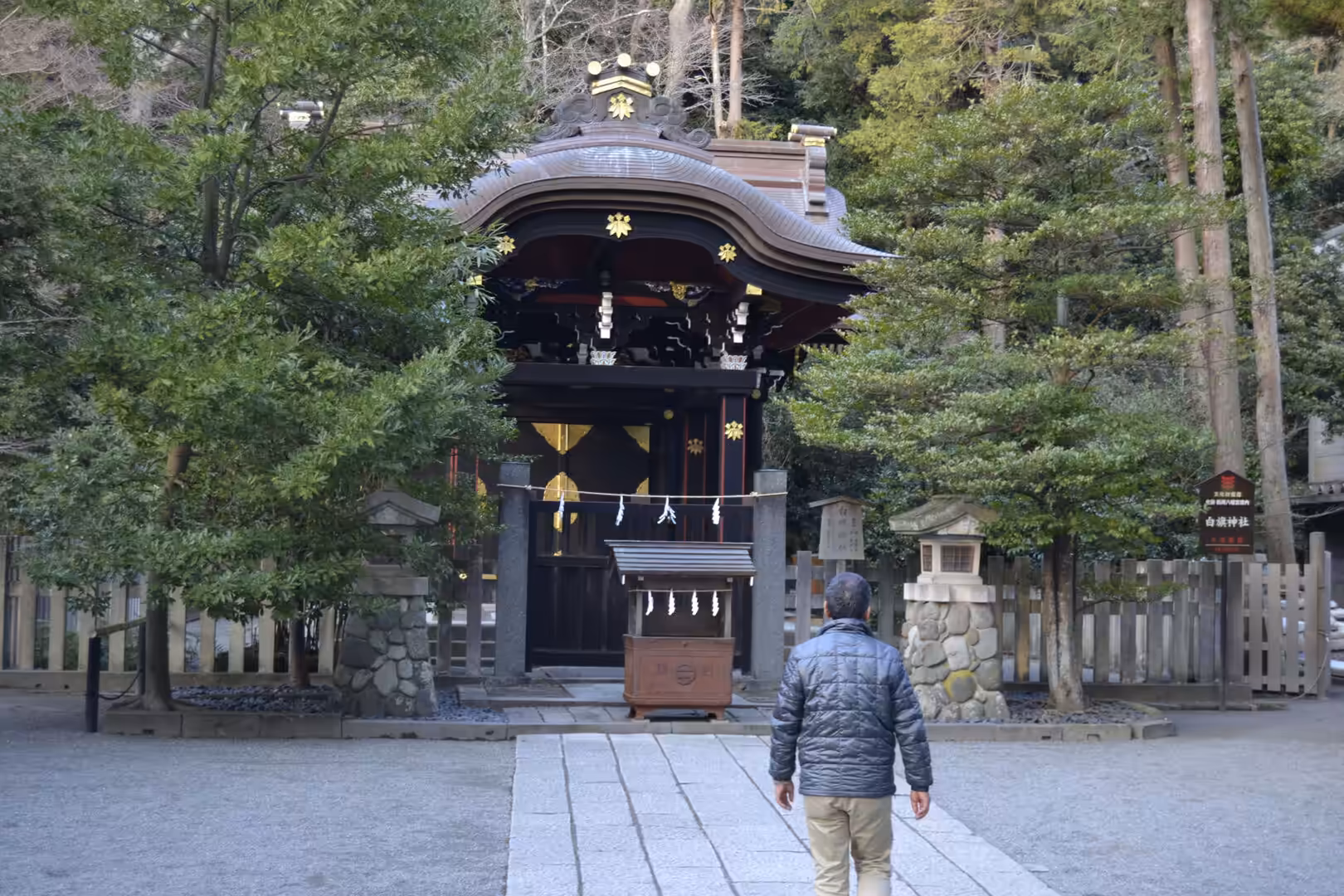 A dark wooden shrine gate with a curved roof stands at the end of a stone path, approached by a lone visitor.