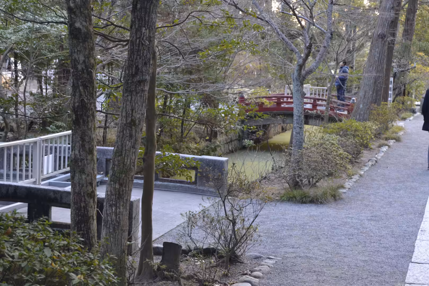 A gravel path runs beside a still pond and stone bridge, with a red footbridge among bare trees in a Kamakura park.