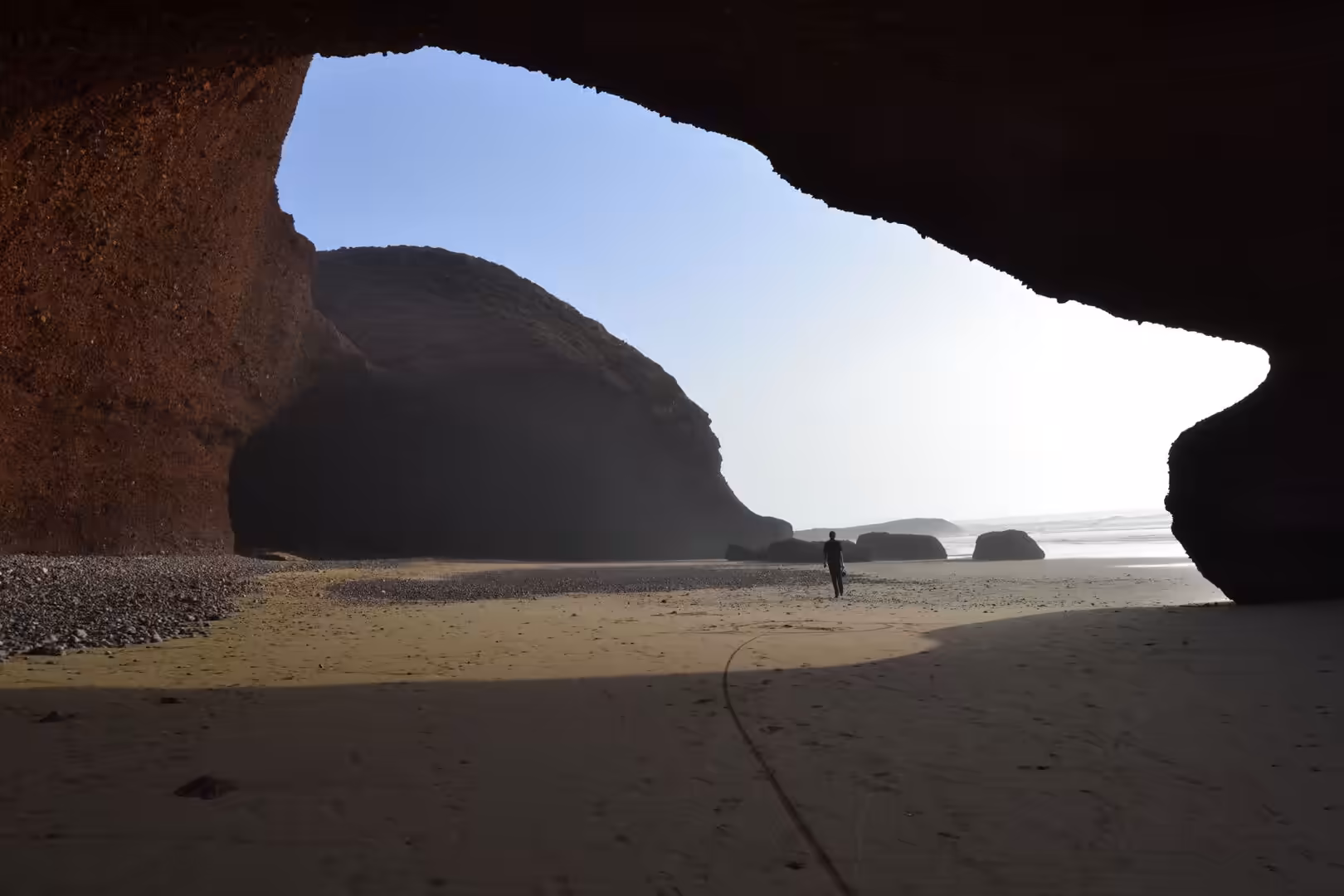 Person walking across a sandy beach beneath a large sea cave opening on the Atlantic coast of Morocco