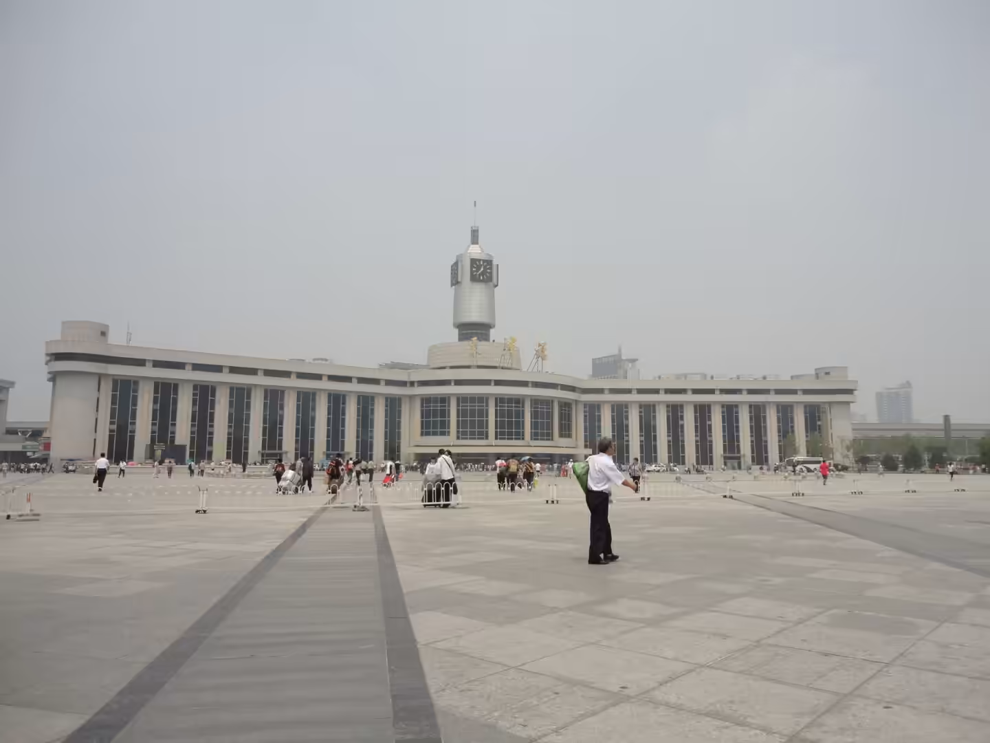 Large railway station building with a central clock tower and wide public square, people walking across the plaza.