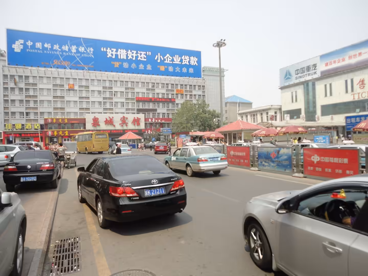 Busy street scene in front of Quancheng Hotel with cars, taxis, buses, and pedestrians under large Chinese billboards