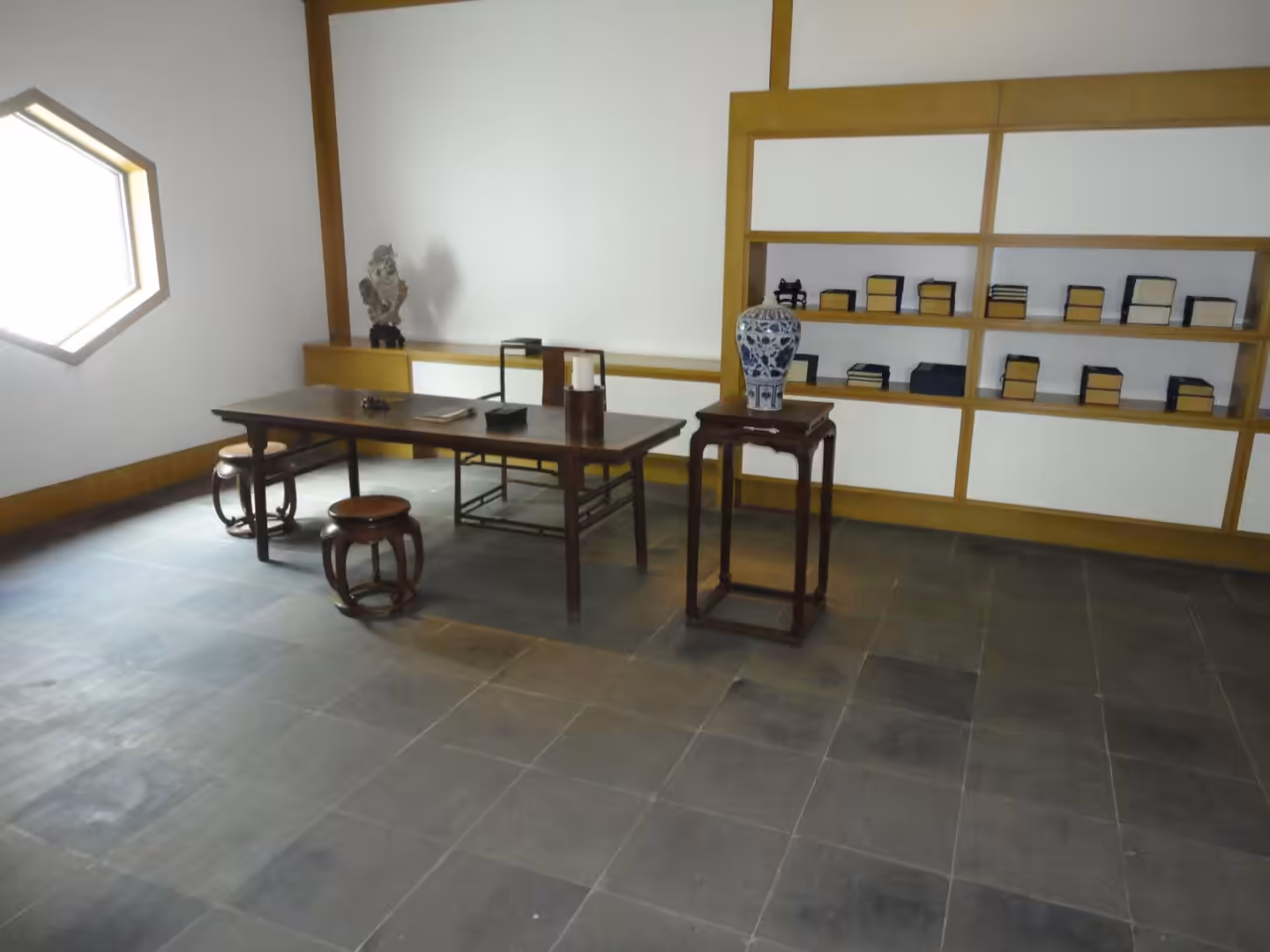 Traditional Chinese scholar’s study with wooden desk, stools, shelves of books, and porcelain vase in a museum interior