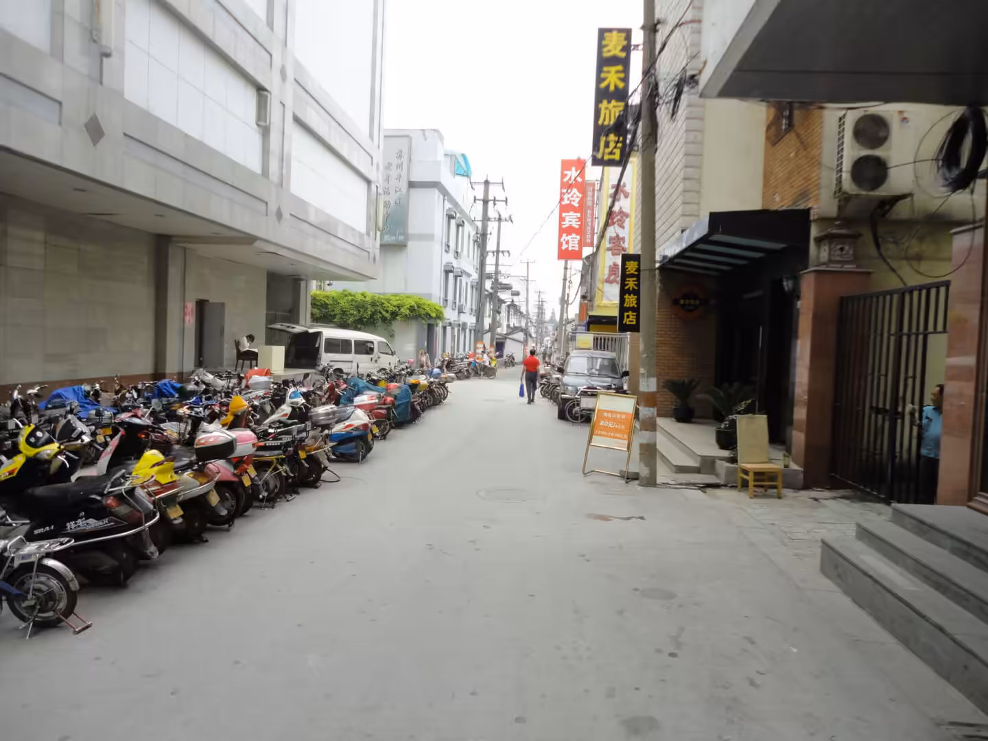 Narrow city street lined with parked scooters, small hotels, and Chinese shop signs, with pedestrians walking along the road.