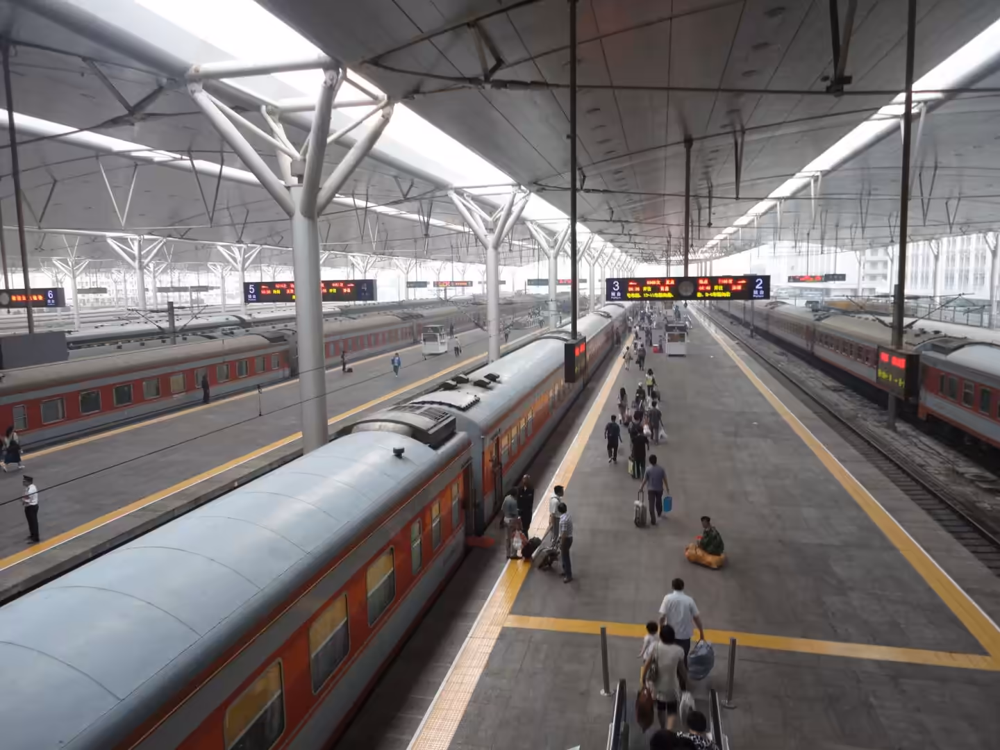 Busy railway platforms with multiple passenger trains, overhead canopy, digital departure boards, and travelers walking with luggage.