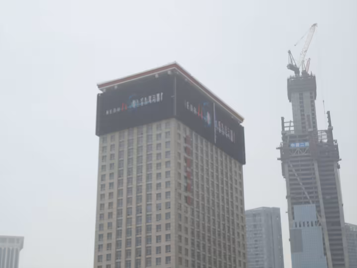 High-rise tower with large digital billboard at the roof and nearby skyscraper under construction in Dalian.