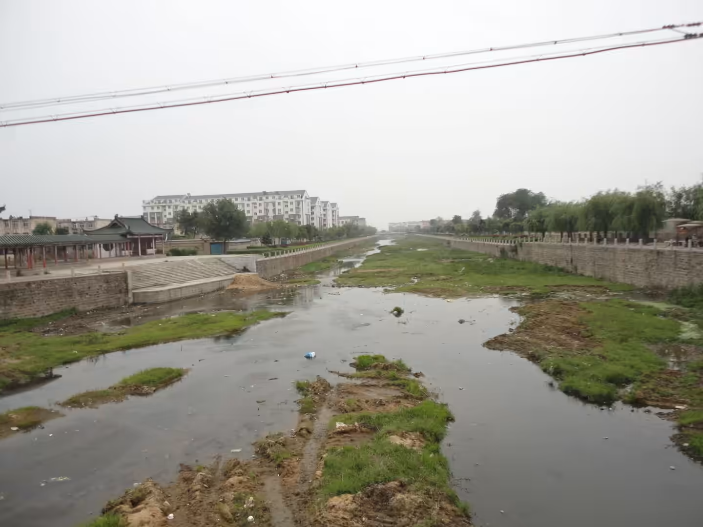 Shallow canal in Xingcheng with stone embankments, patches of grass, low water level, and residential buildings in the distance.