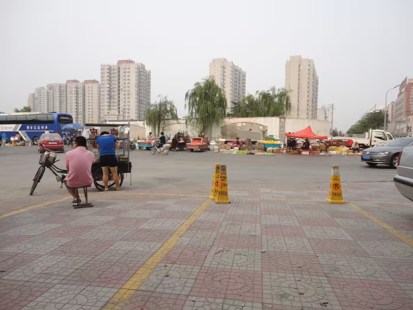 Roadside fruit market in Jinan with vendors, bicycles, passing cars, and high-rise apartments.