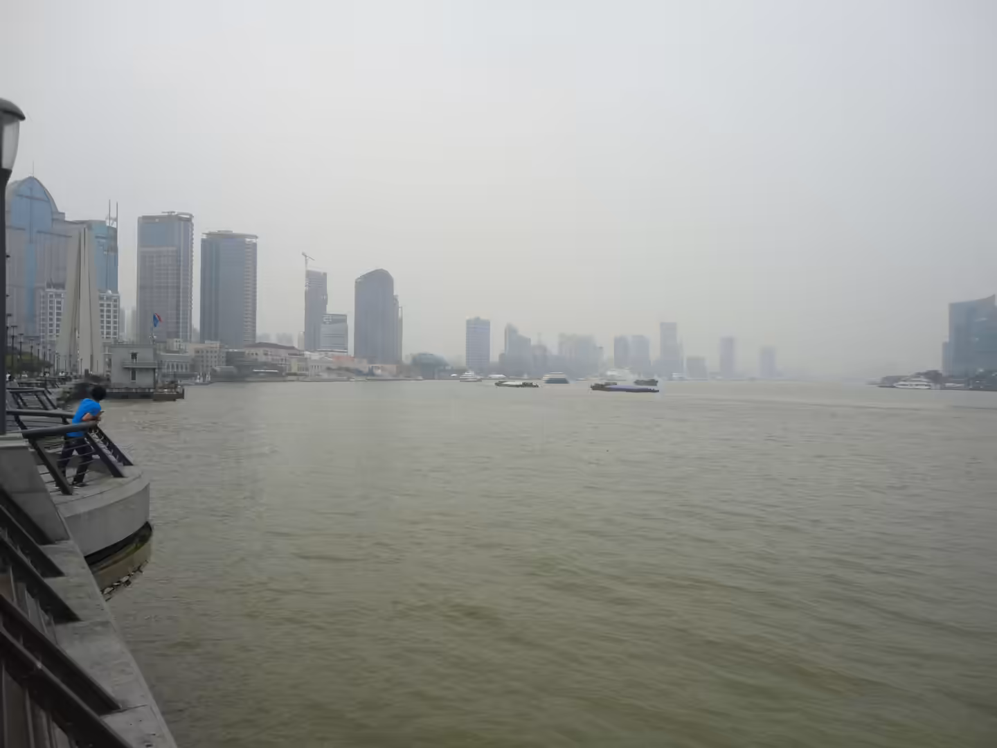 View across the Huangpu River with boats and hazy Shanghai skyline, seen from the Bund riverside promenade.