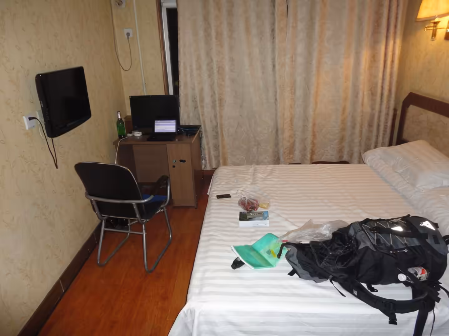 Simple hotel room with single bed, desk, chair, wall-mounted TV, laptop, and travel backpack in Xingcheng.