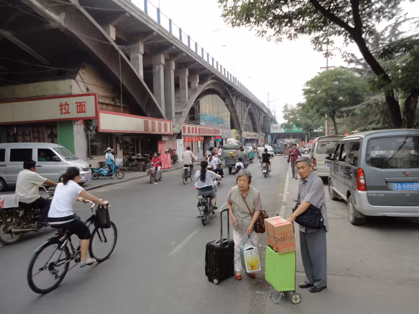 Busy street in Jinan with cyclists, pedestrians, small shops, and traffic beneath an elevated bridge.