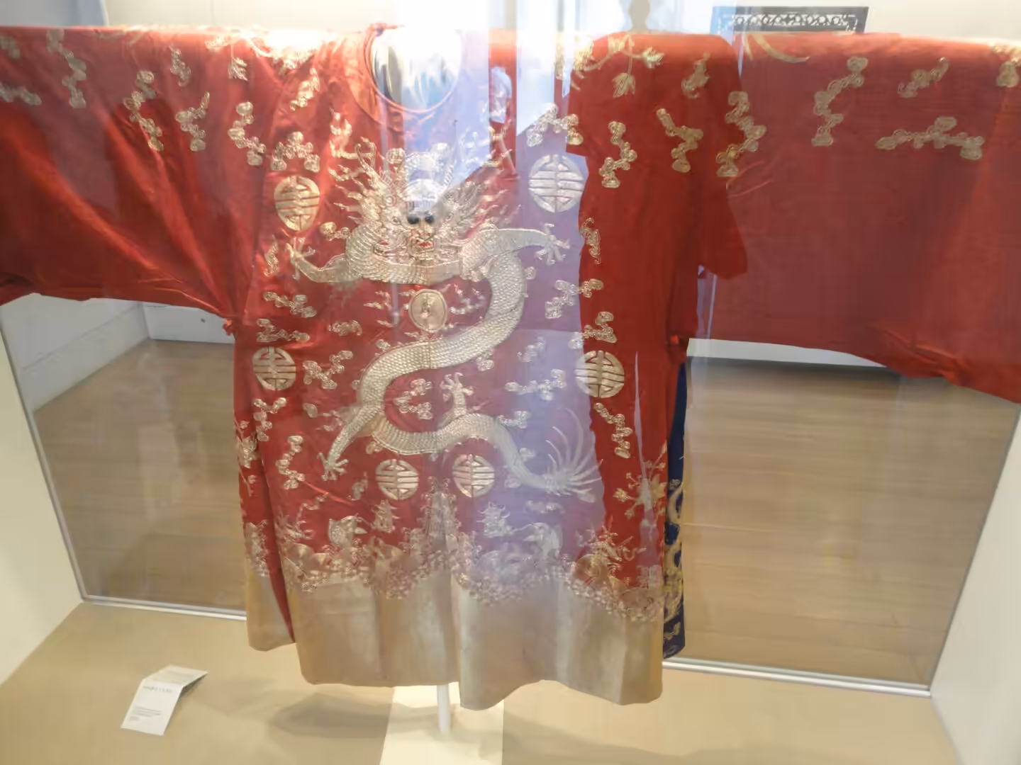 Red silk robe embroidered with silver dragon and traditional Chinese symbols, displayed behind glass