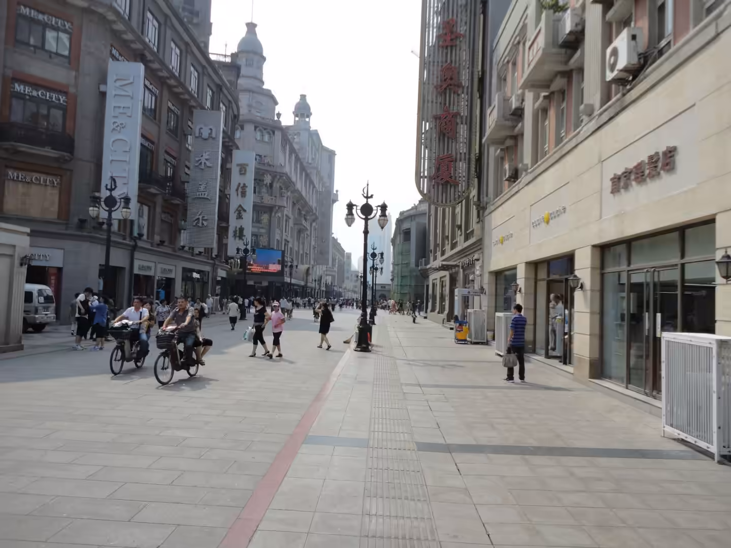 Wide pedestrian shopping street with cyclists, shoppers, European-style façades, and tall commercial buildings.