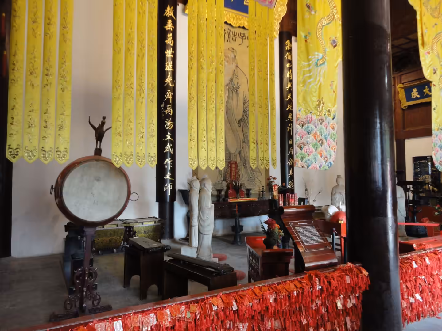 Interior of a Confucian temple hall with yellow hanging banners, statues, drum, and red prayer ribbons