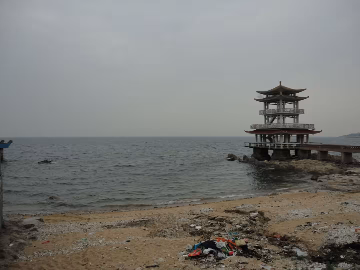 Seaside pavilion on stilts in Xingcheng, standing over rocky shoreline with calm sea and overcast sky.