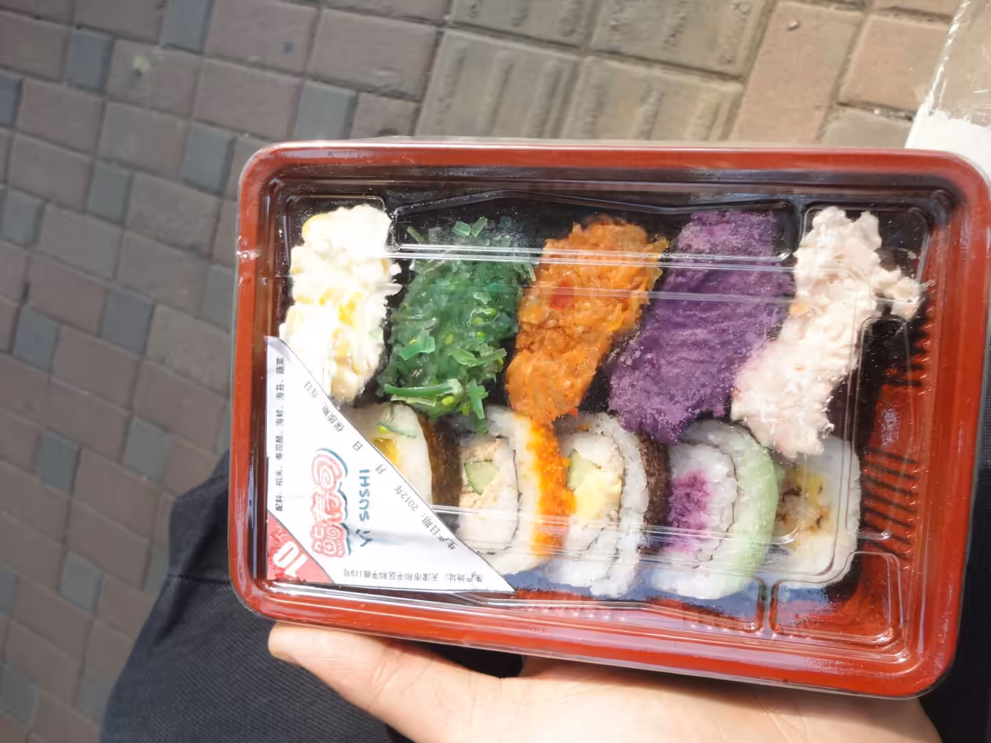 Packaged takeaway sushi set held in hand, with colorful rolls topped with seaweed salad, roe, and mashed vegetables.