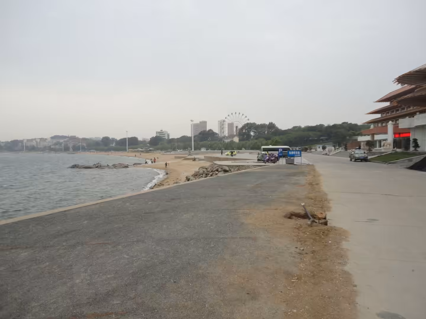 Seafront walkway in Xingcheng with beach, breakwater rocks, calm sea, and distant ferris wheel under a hazy sky.