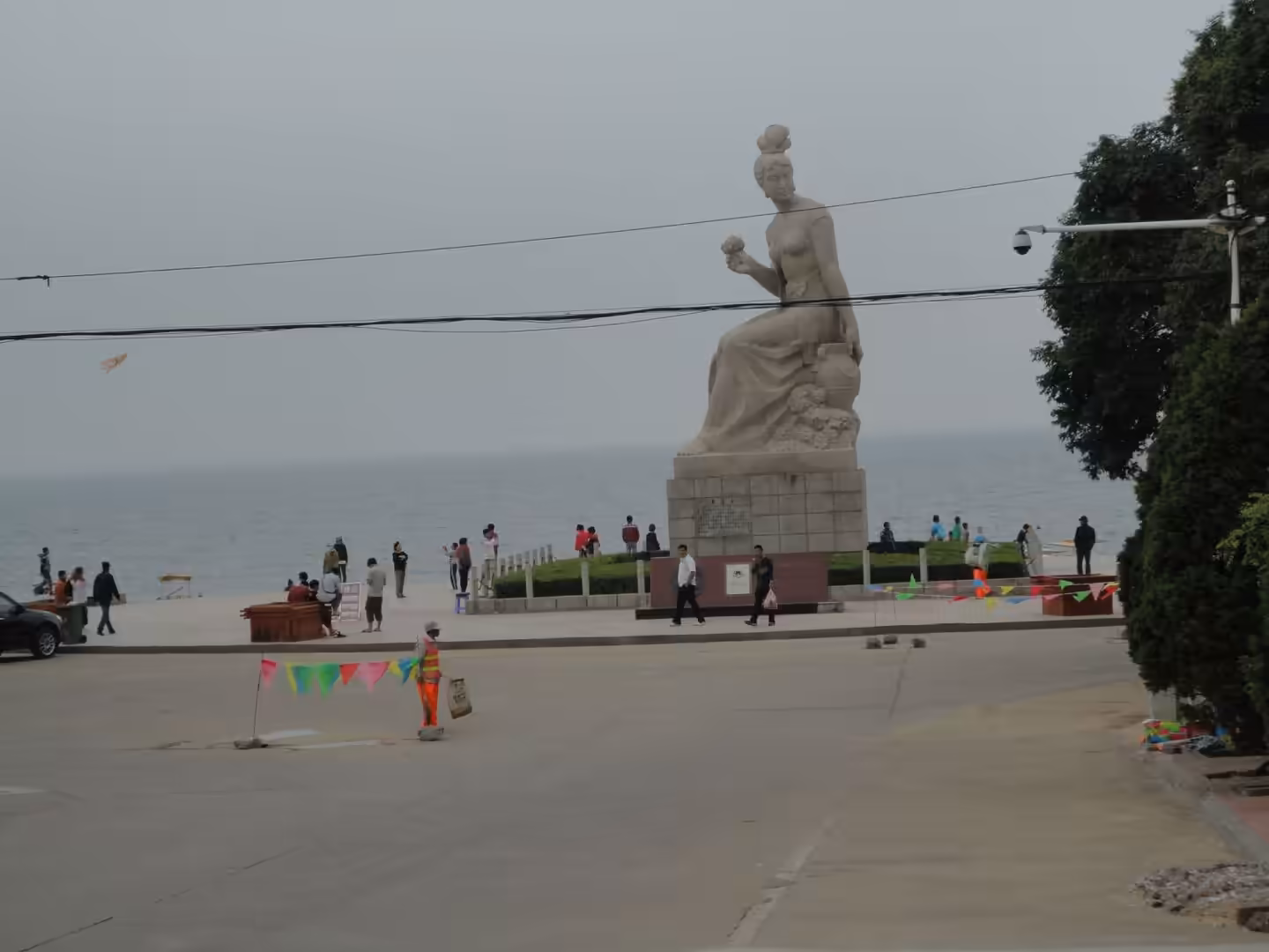 Large seaside statue of a seated woman in Xingcheng, overlooking the sea with visitors walking along the waterfront promenade.