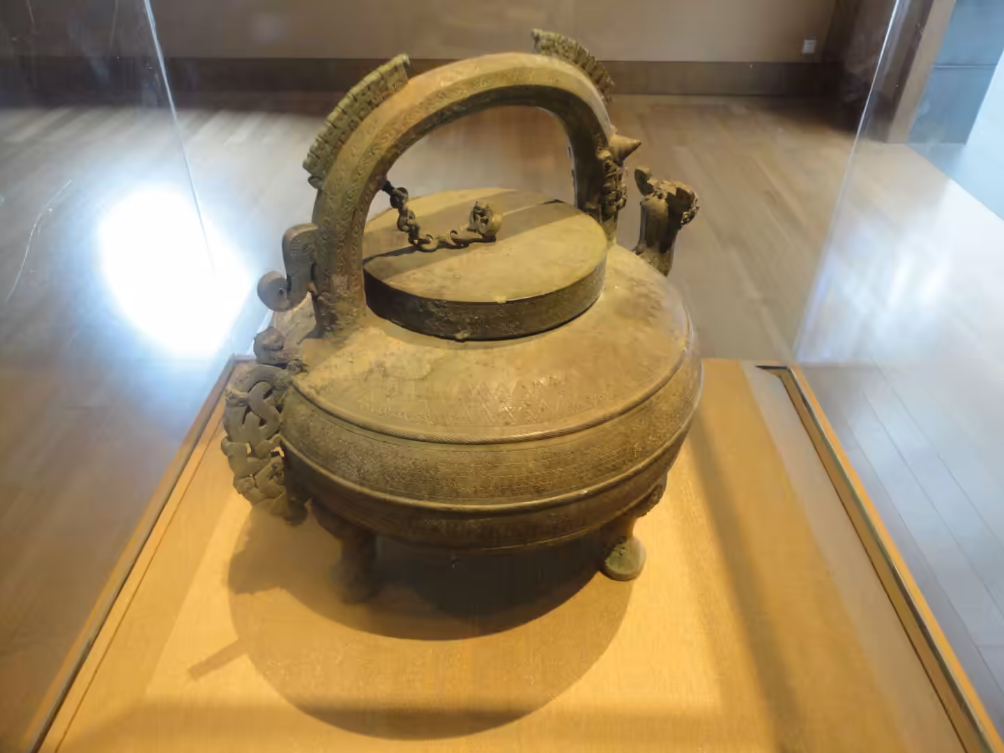 Ancient bronze vessel with arched handle and decorative animal motifs, displayed in a glass case