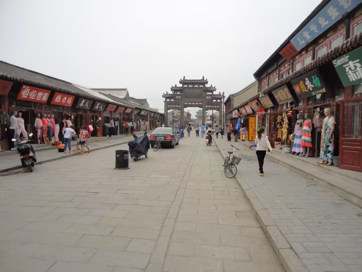 Wide stone-paved market street in Xingcheng Ancient City with traditional shopfronts, pedestrians, scooters, and a historic gate in the distance.