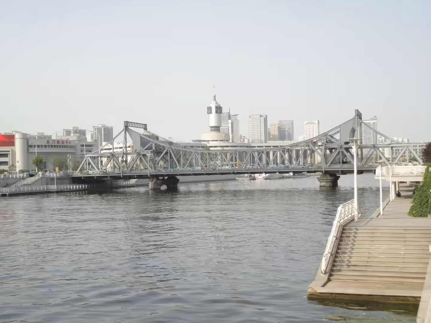 Steel truss bridge crossing the Haihe River in Tianjin, with calm water, riverside steps, and modern city buildings behind.