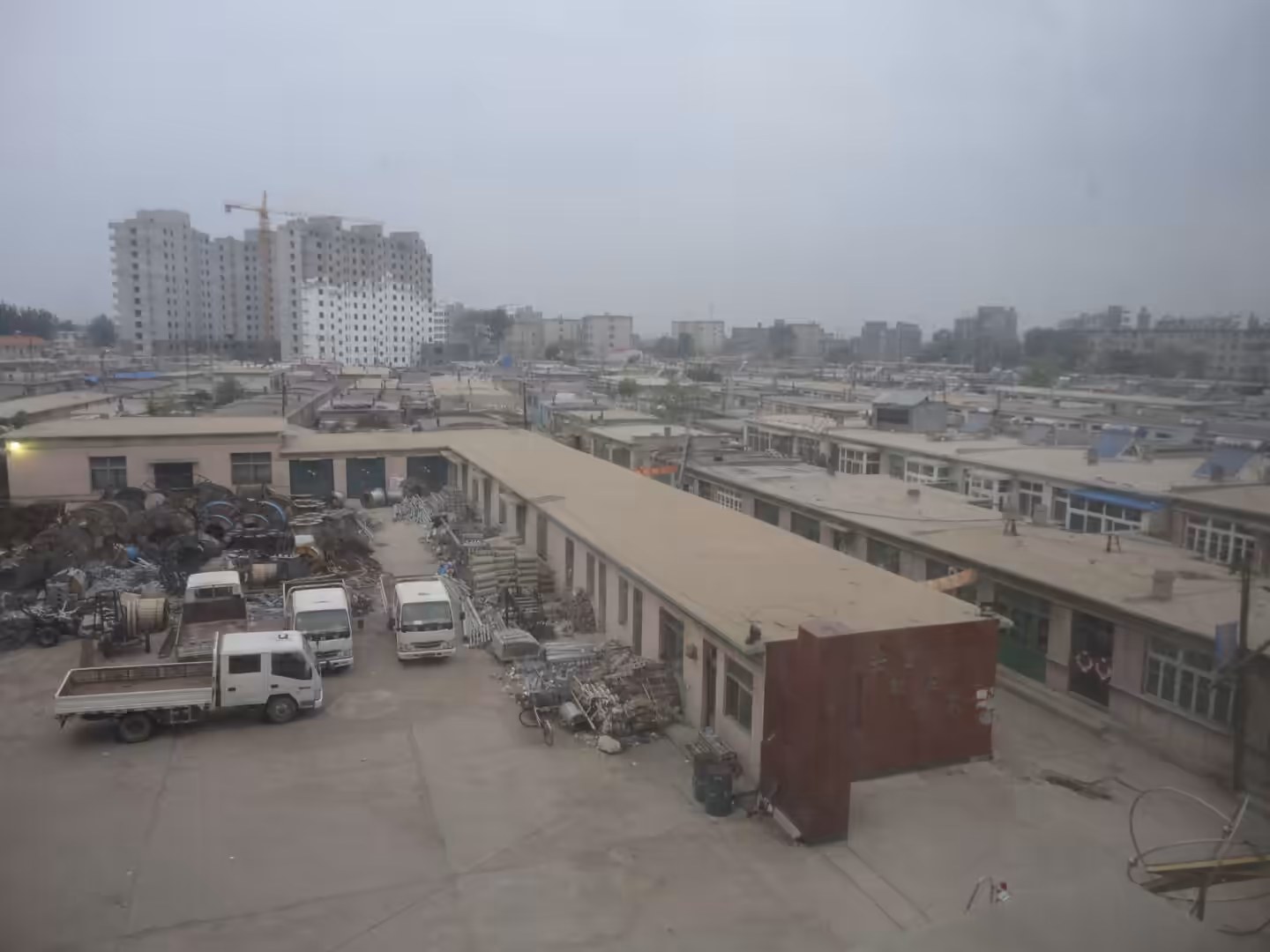 Industrial workshop yard in Xingcheng with low factory buildings, stacked metal reels, parked trucks, and distant apartment blocks under construction under a hazy sky.