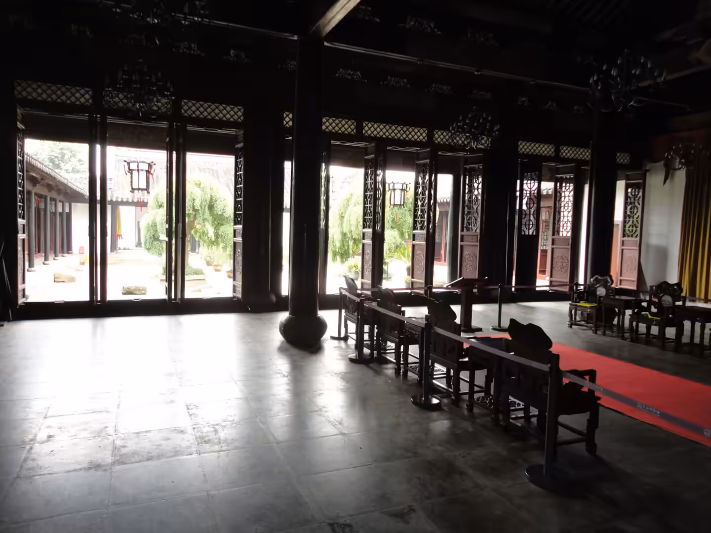 Dimly lit ceremonial hall with dark wooden columns, carved chairs, red carpet, and open courtyard doors