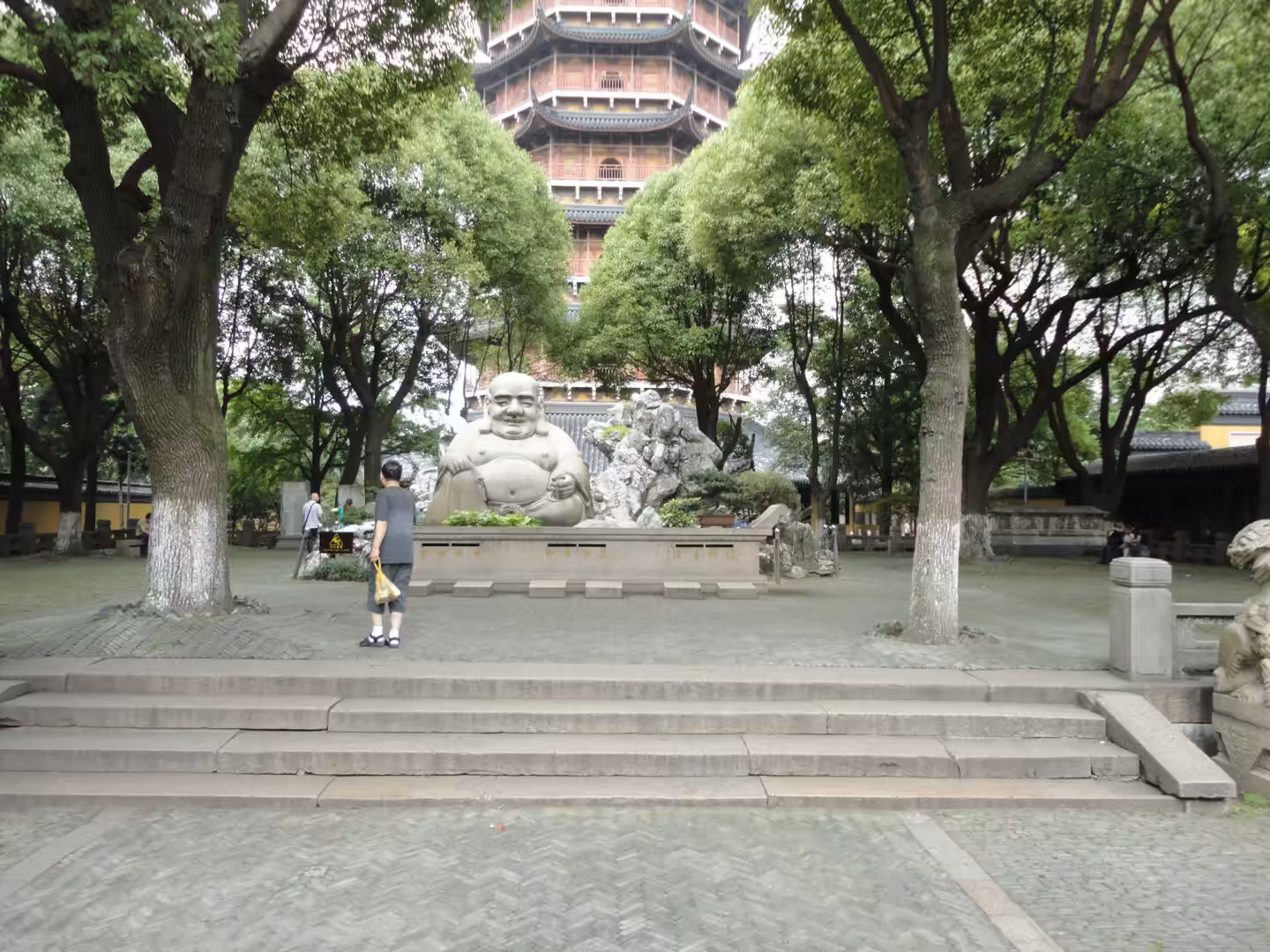 Smiling Buddha statue in a tree-lined temple courtyard with a tall pagoda rising behind