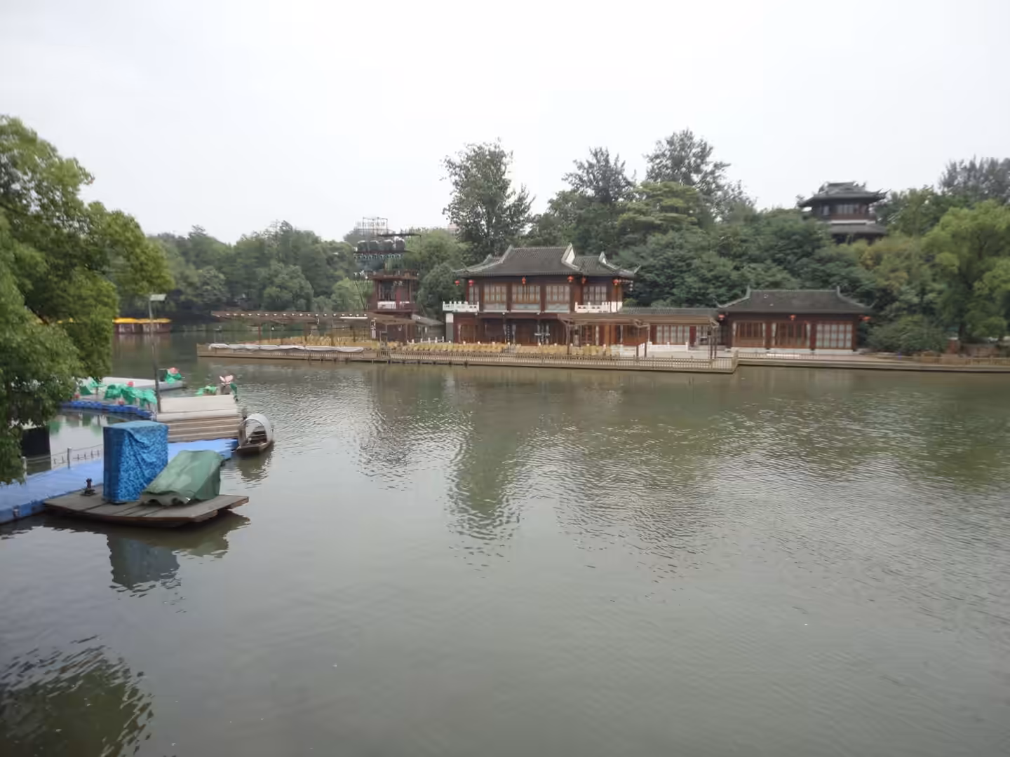 Traditional Chinese buildings along a calm river with boats, trees, and wooden pavilions