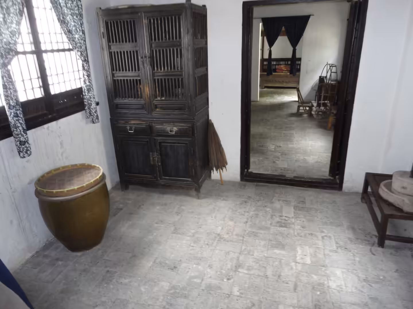 Interior of a traditional silk worker’s home with wooden cabinet, ceramic jar, and simple furnishings on a stone floor.