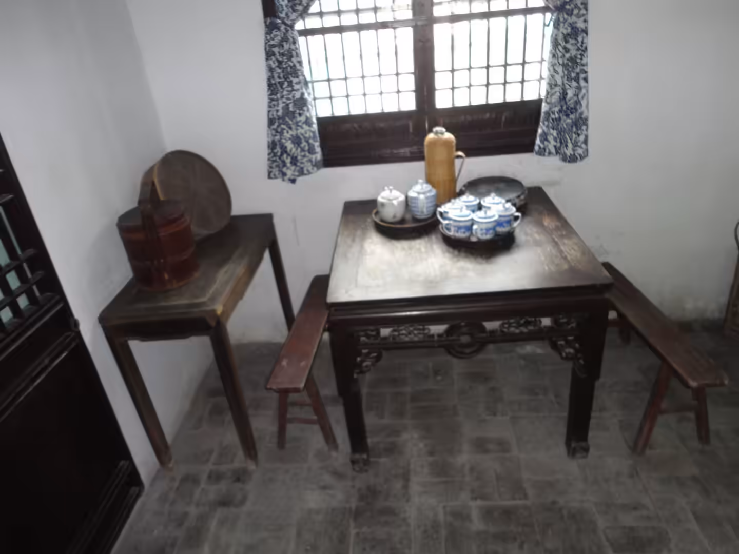 Small wooden table with tea set, benches, and a side table beneath a lattice window in a traditional interior.