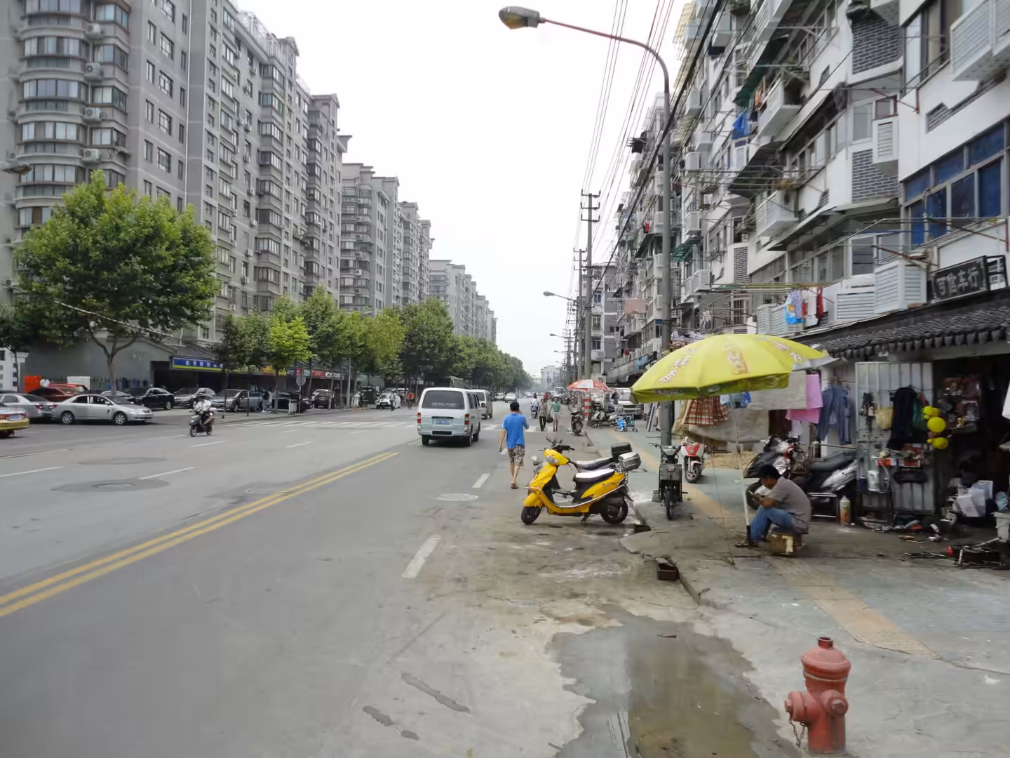 Wide urban street with apartment blocks, scooters, small sidewalk shops, and pedestrians