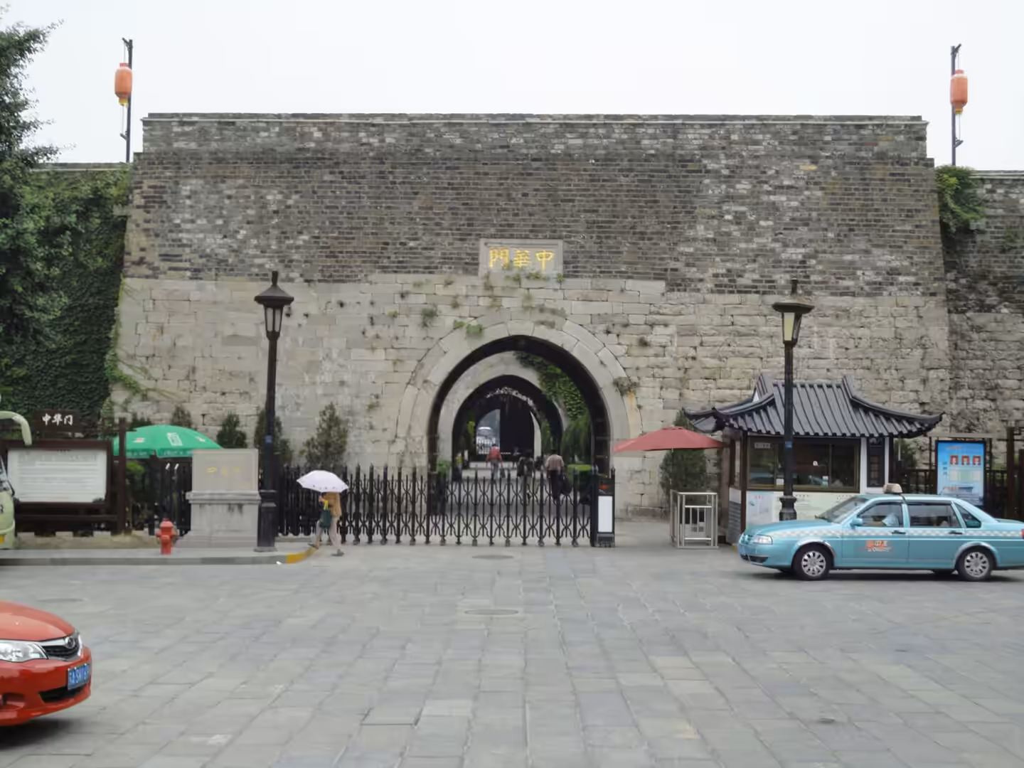 Stone city gate with an arched entrance, pedestrians walking through, and taxis parked in front
