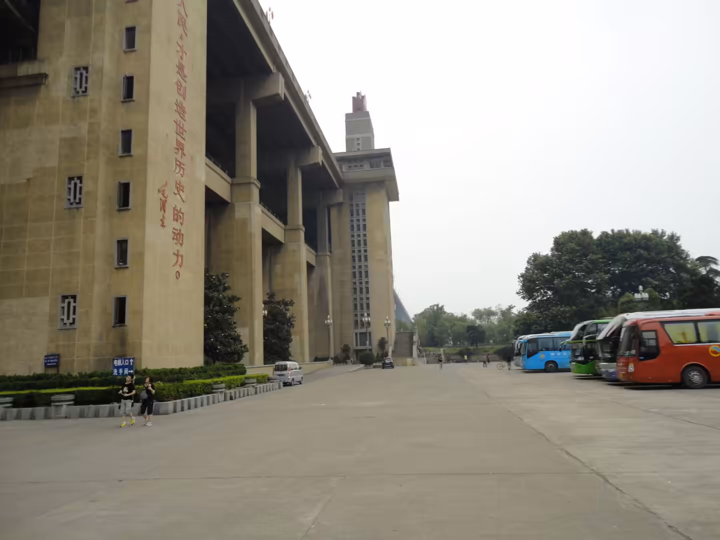 Large concrete riverside building with tall pillars, open plaza, and parked tour buses