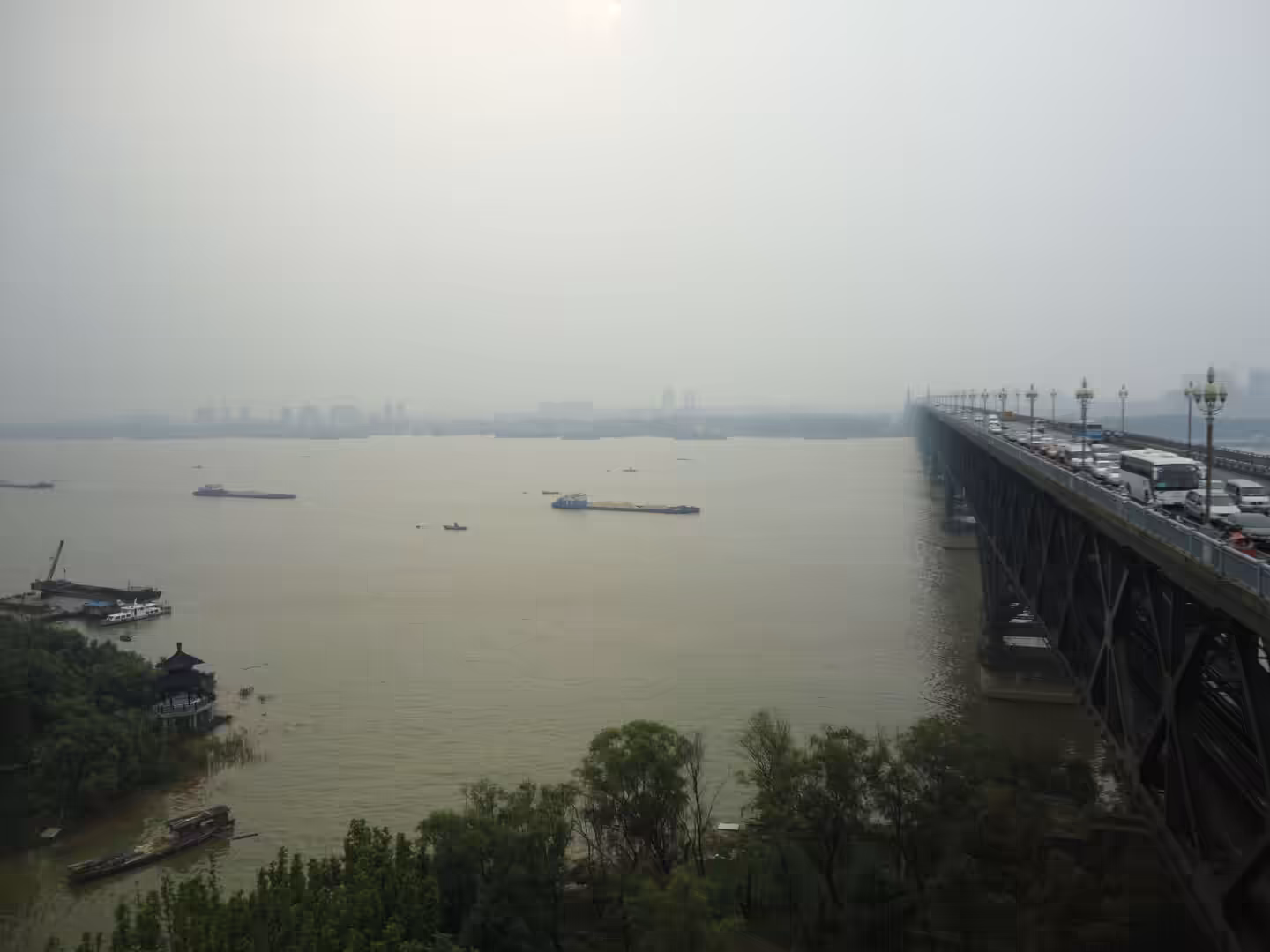 Yangtze River flowing beneath the Nanjing Yangtze River Bridge, with cargo boats on the water and heavy traffic crossing the steel structure under a hazy sky.