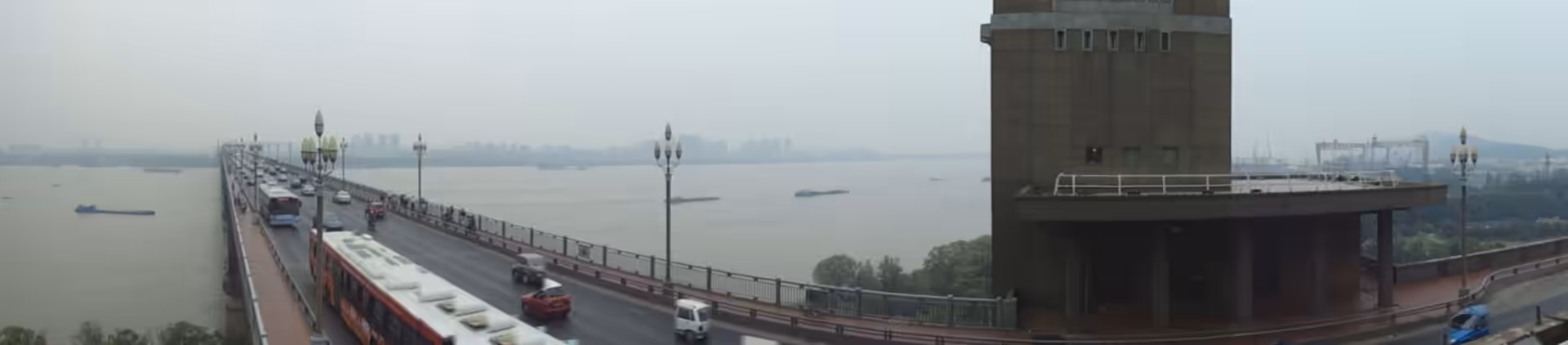 Traffic crosses the Nanjing Yangtze River Bridge with buses, cars, and pedestrians above the wide Yangtze River under a hazy sky.