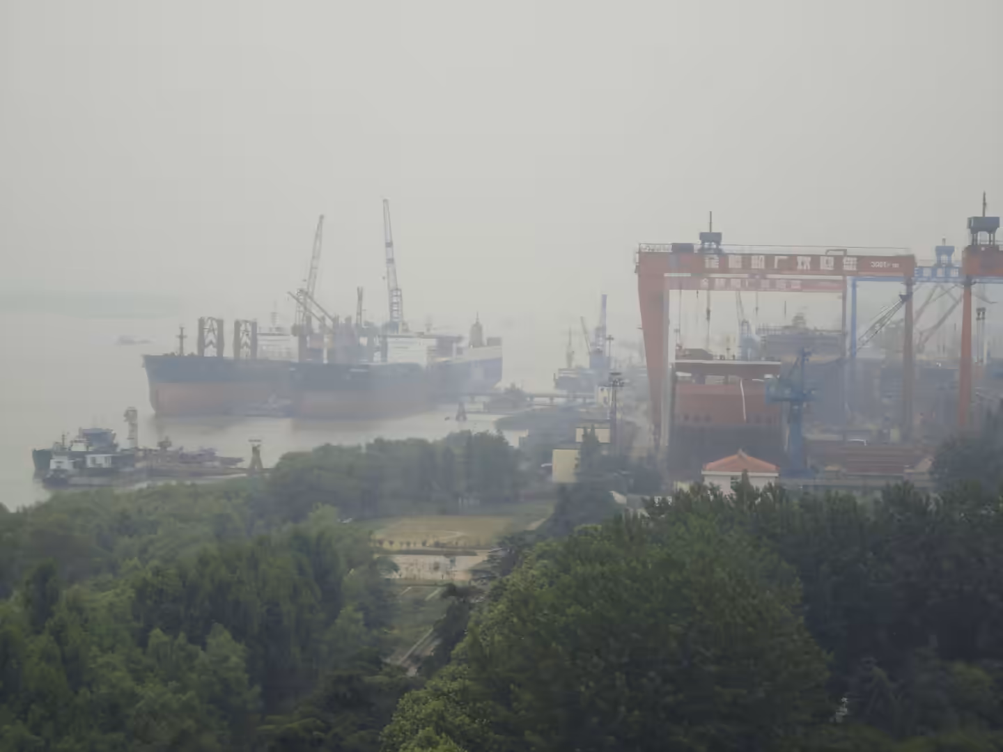 Shipyard cranes and large vessels along the Yangtze River seen through heavy haze, with green riverside park in the foreground.