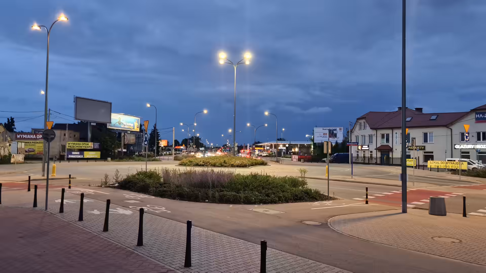 A modern roundabout in Warsaw at dusk with illuminated streetlights under dramatic storm clouds.