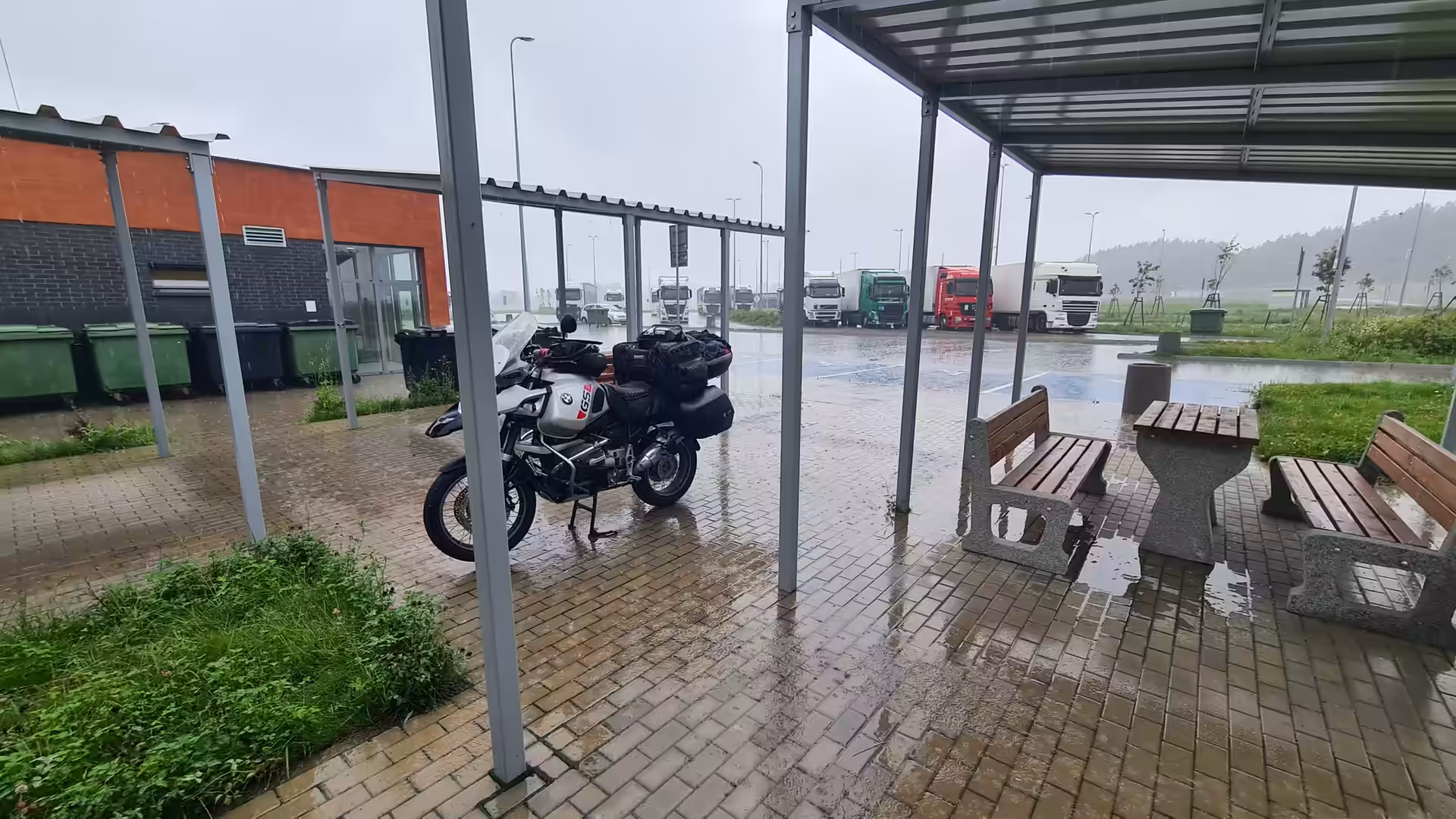 A BMW GS Adventure motorcycle parked under a shelter during heavy rain at a highway rest area with trucks in the background.