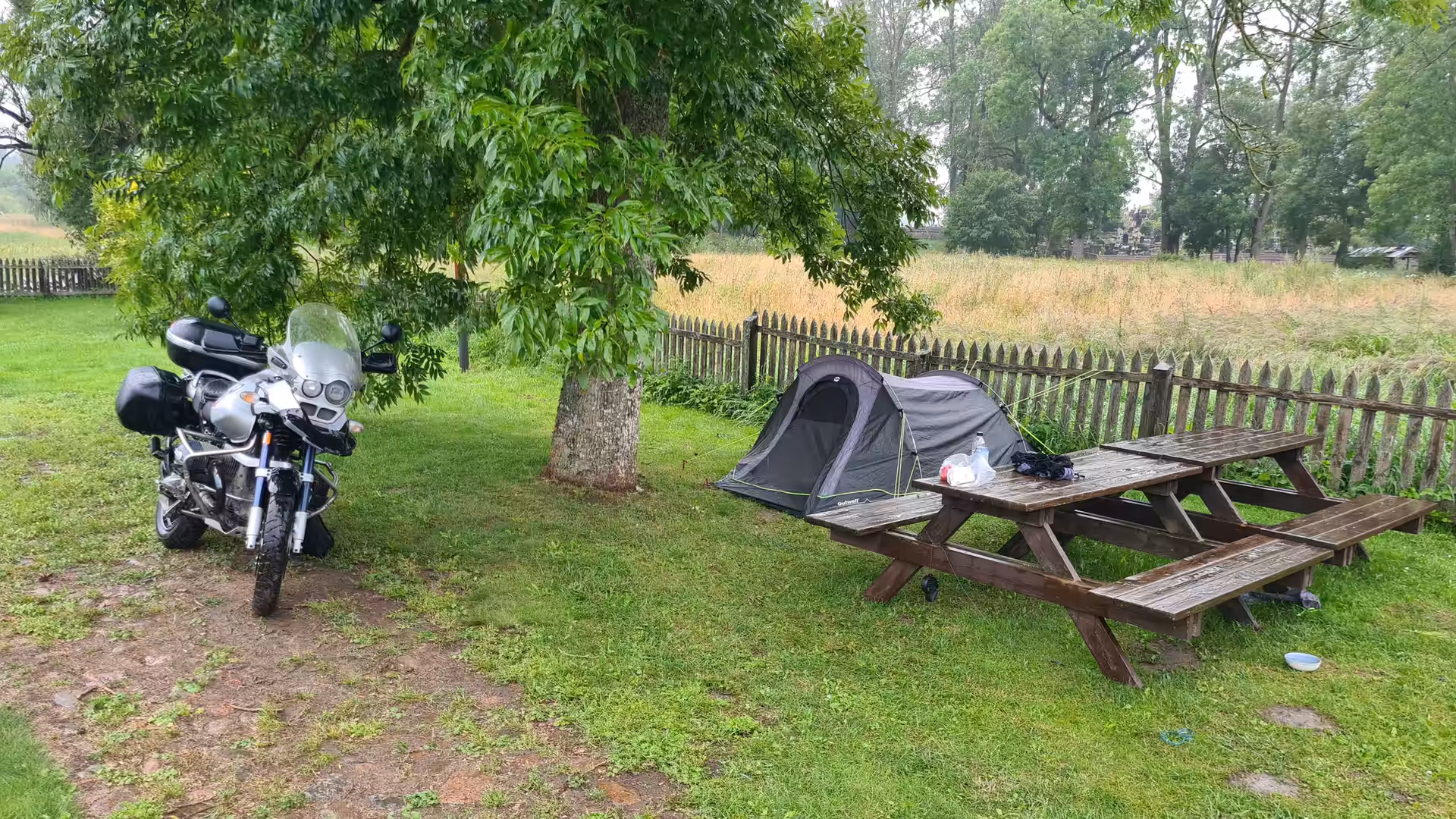 A BMW GS Adventure parked beside a small tent at a quiet campsite under a large tree during a rainy day.