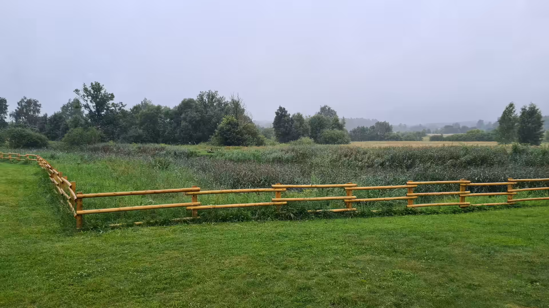 A peaceful rural landscape with green meadows, wooden fencing, and trees under a gray, rainy sky.