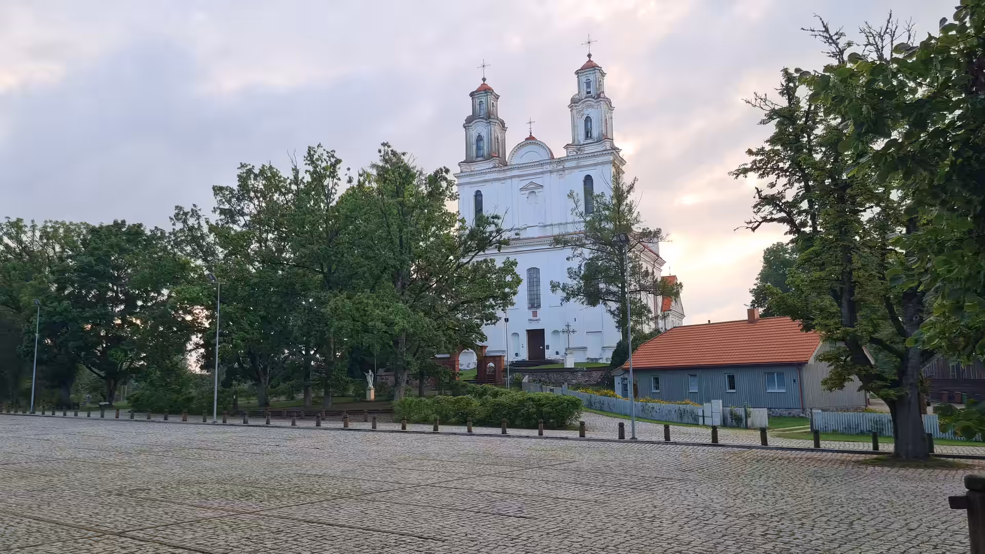 A historic white church with twin towers overlooking a cobblestone square in Lithuania during sunset.